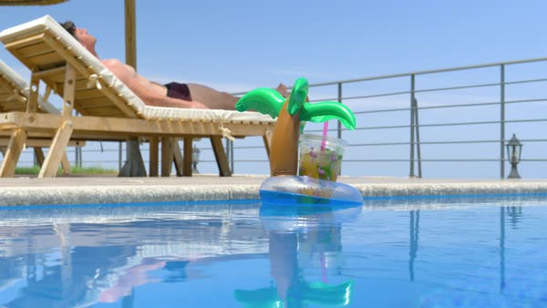 Inflatable Drink Holder With Plastic Mug Full Of Orange Juice Floating In The Pool Adult Male Lying On A Deck Chair Under Umbrellas In The Background