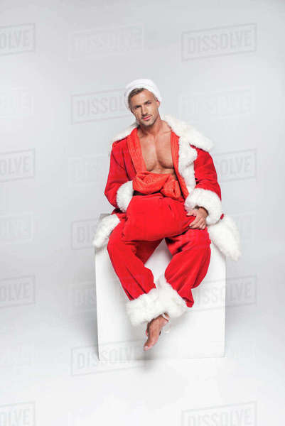 Sexy man in santa costume sitting with red bag and looking at camera on grey - Royalty-free Stock Photo | Dissolve Sexy man in santa costume sitting with red bag and looking at camera on grey - Royalty-free Stock Photo | Dissolve