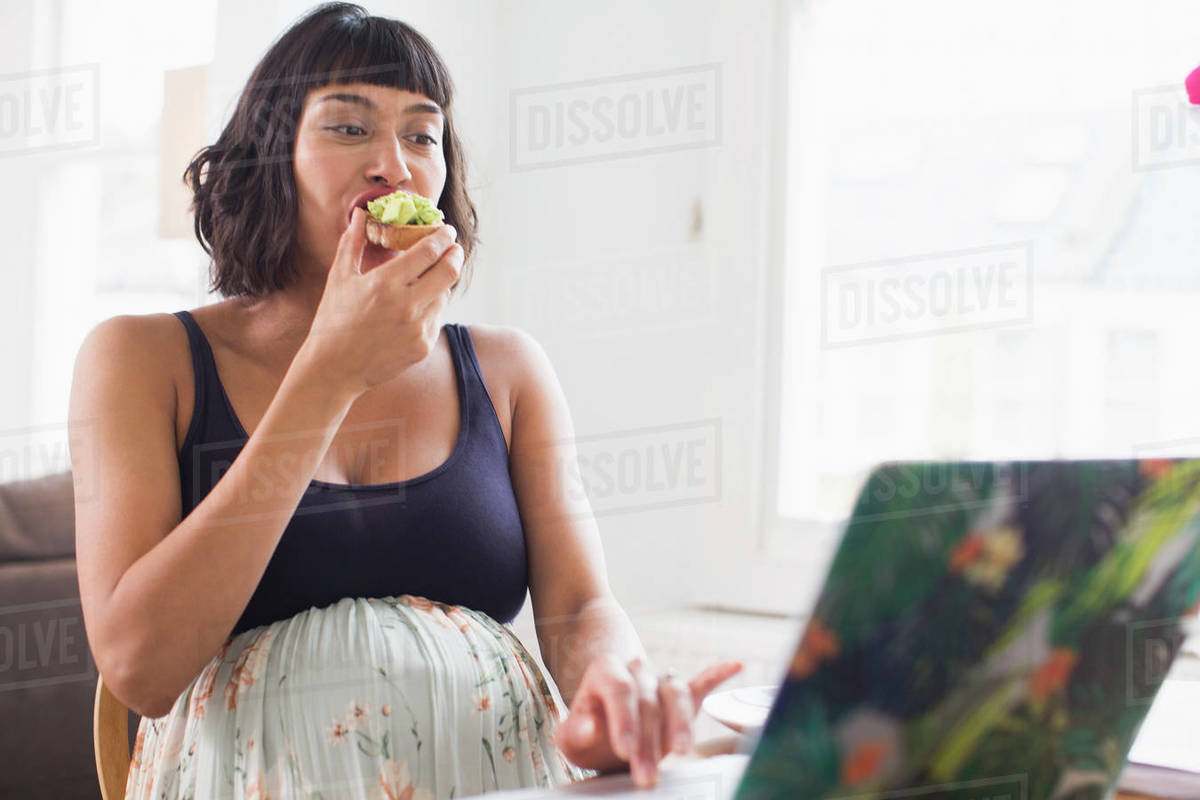 Pregnant woman eating avocado toast at laptop Stock Photo Dissolve