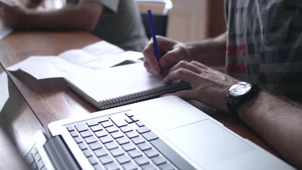 close up of man writing in notebook with laptop computer nearby