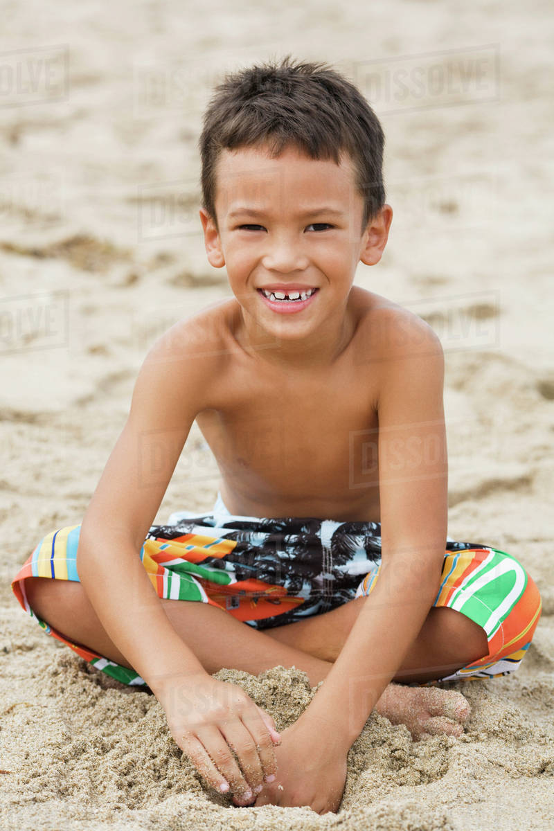 Mixed race boy in swim trunks sitting on beach Stock Photo Dissolve