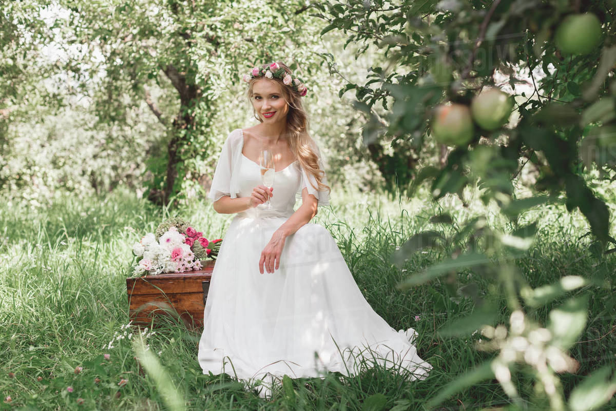 beautiful young bride holding glass of champagne and smiling at camera while sitting on vintage chest in garden - Royalty-free Stock Photo | Dissolve beautiful young bride holding glass of champagne and smiling at camera while sitting on vintage chest in garden - Royalty-free Stock Photo | Dissolve