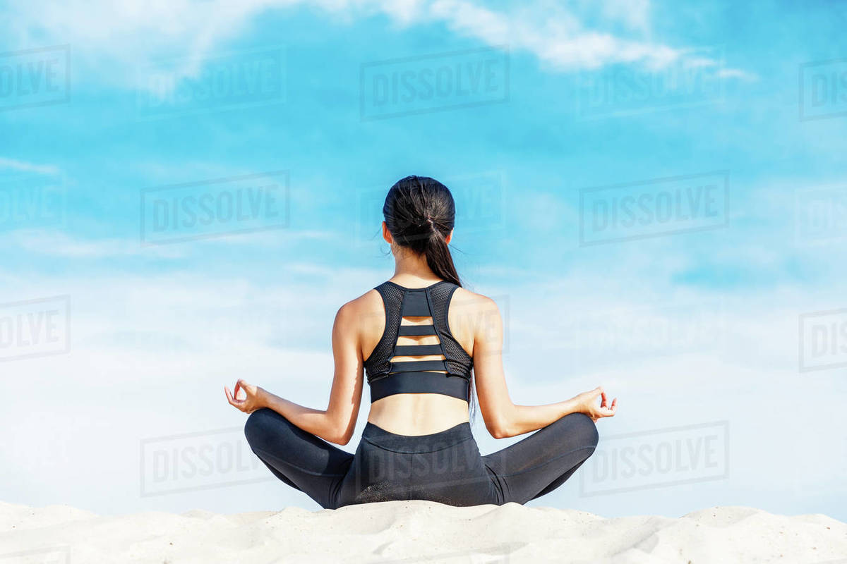 rear view of woman meditating in lotus yoga pose on sand against cloudy ...