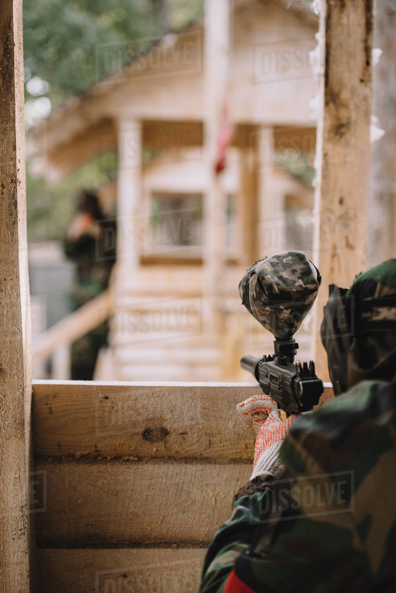 Partial view of paintball player in camouflage uniform aiming by paintball gun from wooden tower