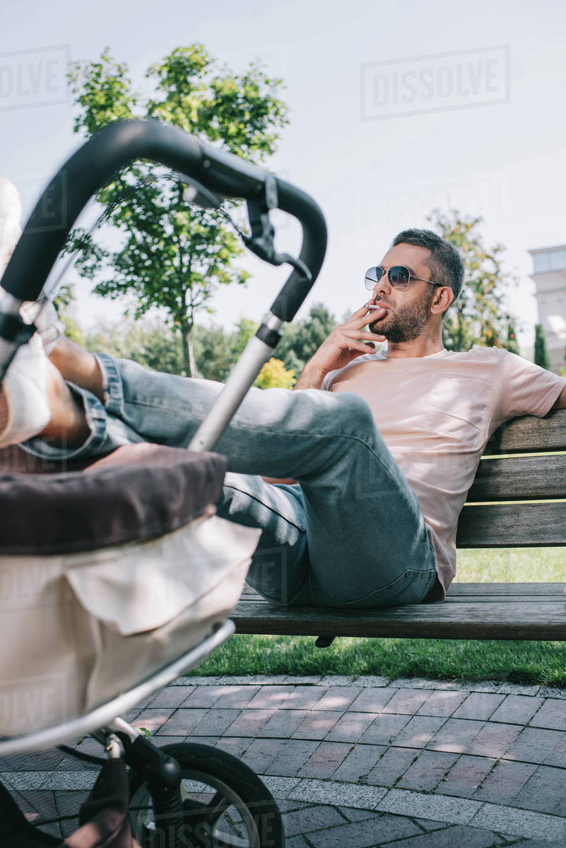 dad smoking cigarette Father smoking cigarette on bench with legs on baby carriage in park -  Royalty-free Stock Photo