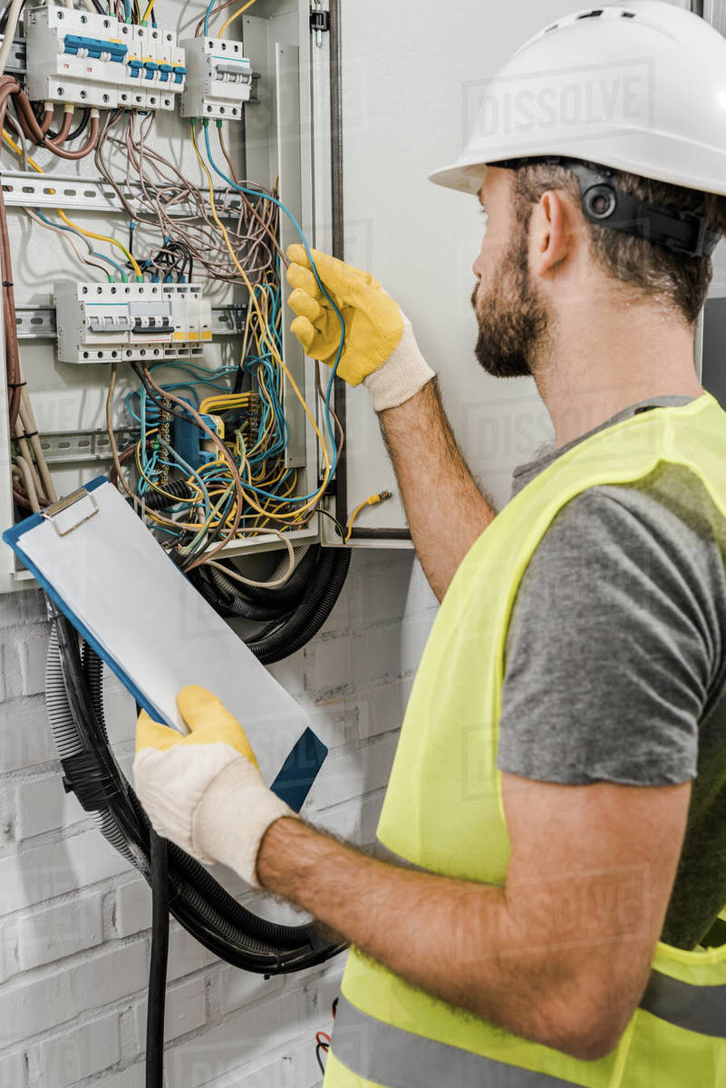 Electrician holding clipboard and checking wires in electrical box in