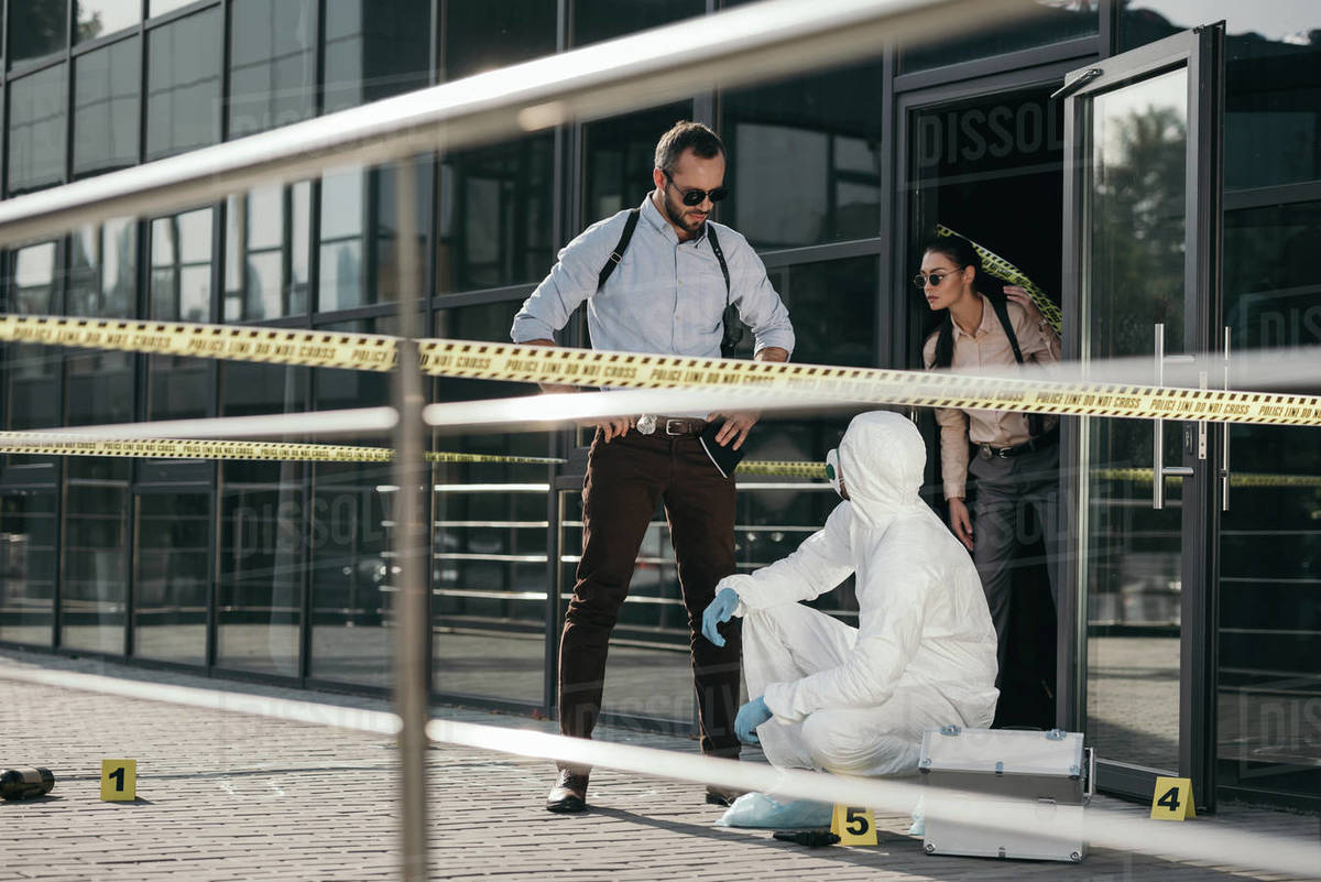 Detectives and criminologist in protective suit and latex gloves communicating at crime scene