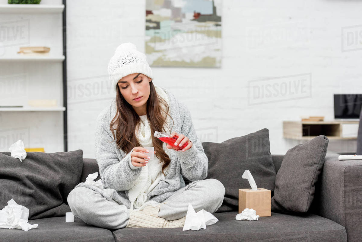 Diseased young woman in warm clothes pouring red cough syrup while