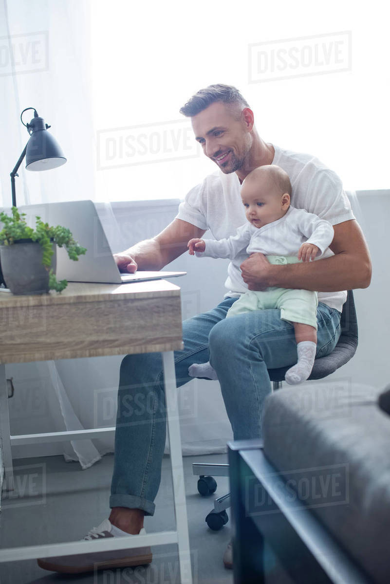 Happy father sitting on chair, holding baby and using laptop at home