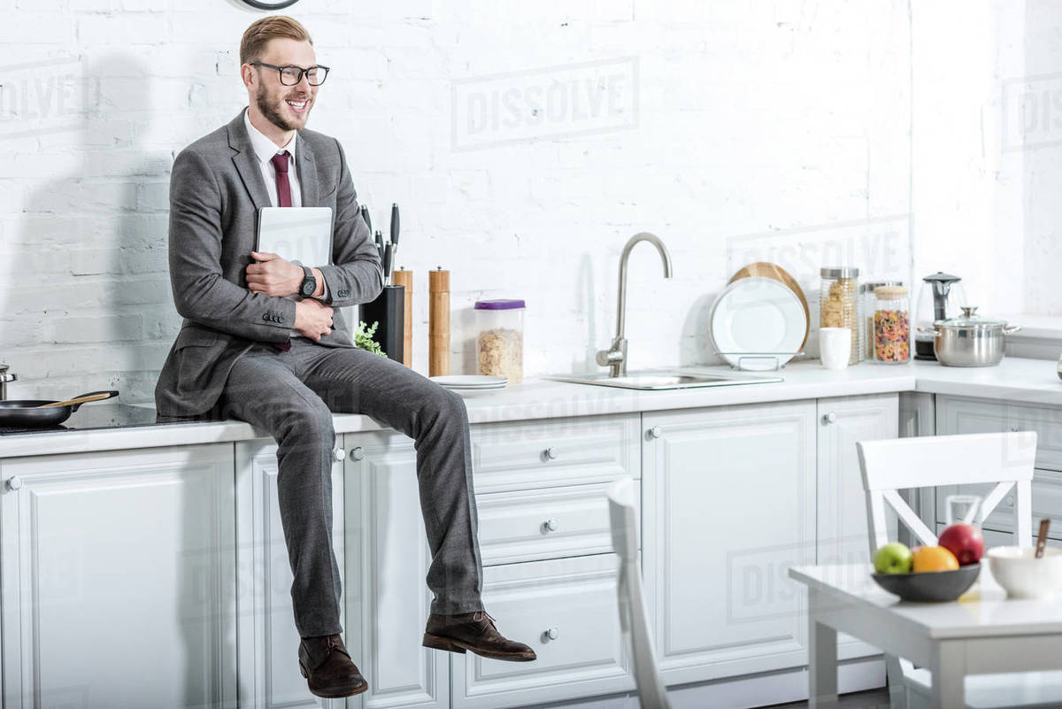 businessman sitting on countertop and holding laptop in kitchen at home
