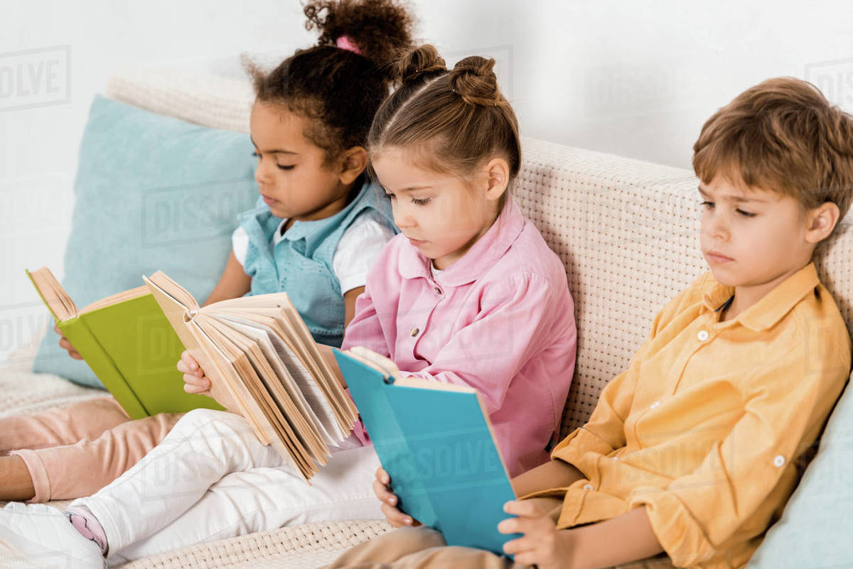 Adorable multiethnic children sitting on sofa and reading books ...