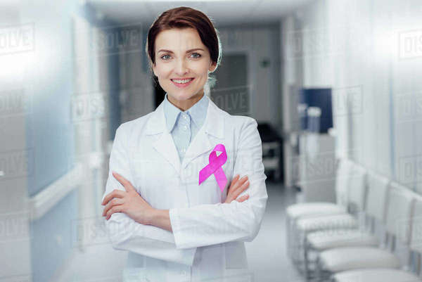 Smiling female doctor with pink ribbon and arms crossed in hospital ...