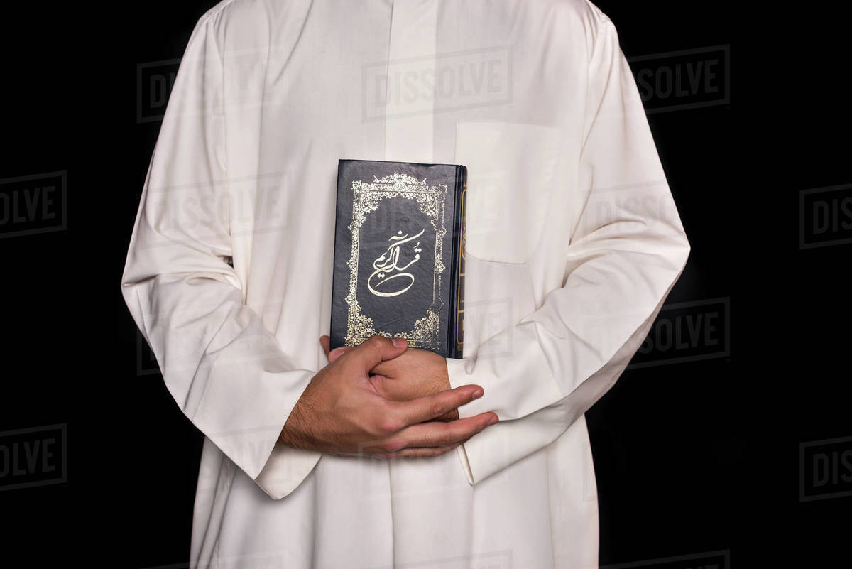 cropped shot of man in muslim clothing holding quran isolated on black Stock Photo Dissolve