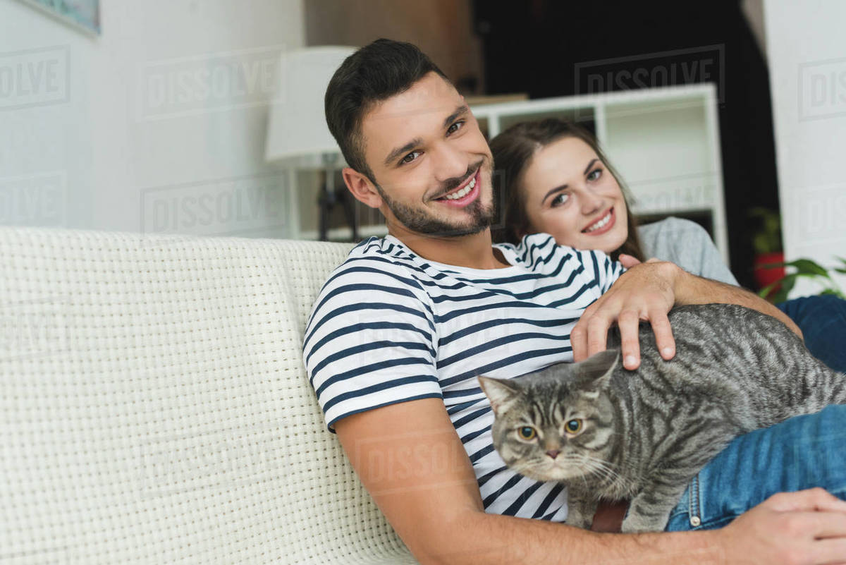 Beautiful Young Couple With Adorable Tabby Cat Sitting On Couch