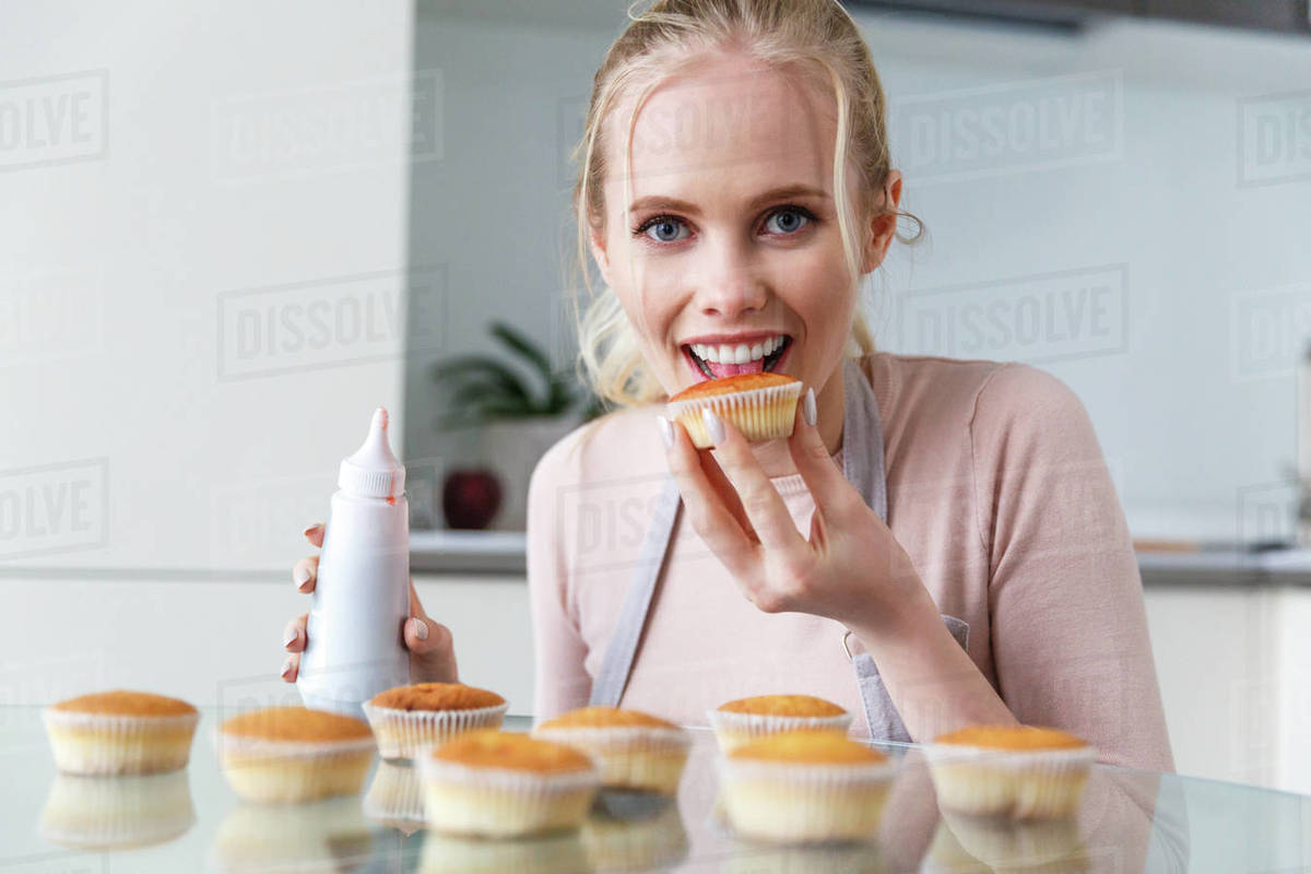 Beautiful young woman eating muffin and looking at camera while cooking