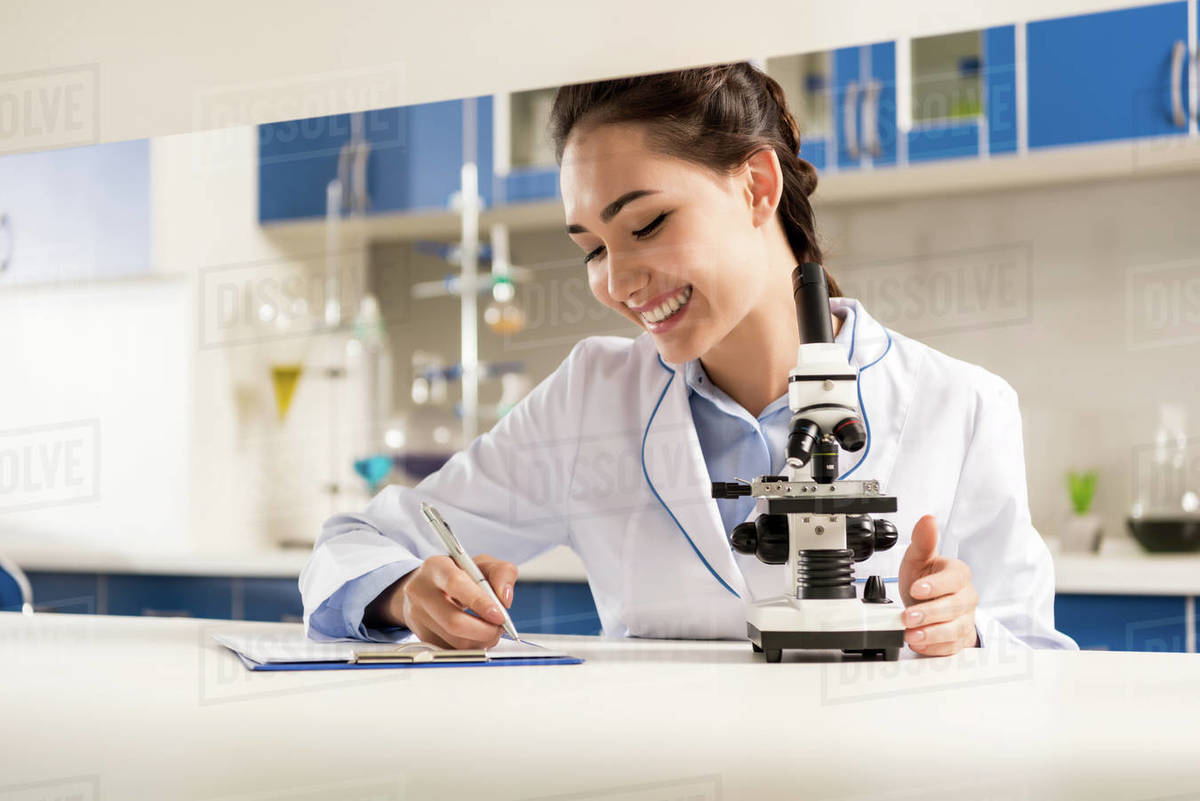 Young smiling lab technician taking notes after doing microscope sample ...