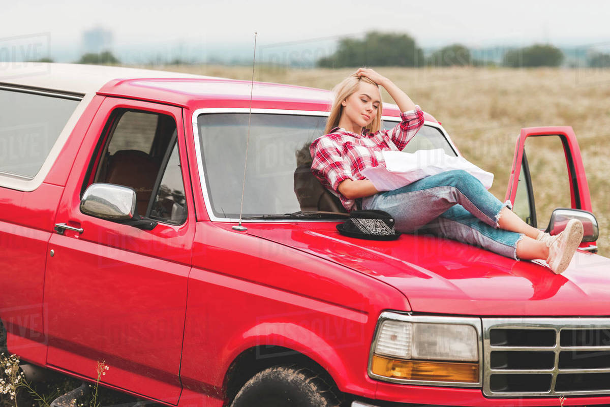beautiful young woman relaxing on hood of car in field Stock Photo Dissolve