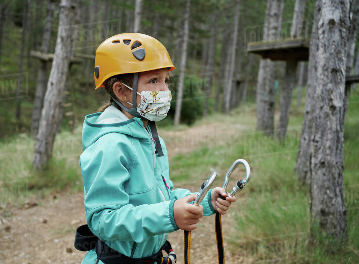 Girl in helmet and fabric mask holding carabiners with ropes and looking away while preparing to climb obstacles in adventure park in forest during pandemic Royalty-free stock photo