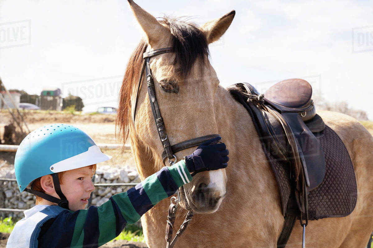 Boy in riding hat and boot walking along paddock with dun horse during