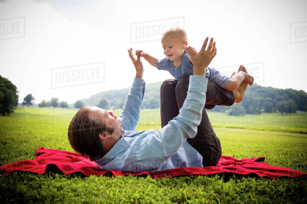 Mid adult man lying on back balancing baby son in rural field Stock