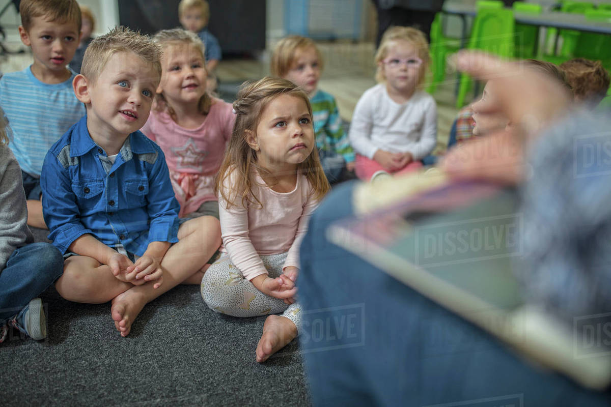 Young children sitting on carpet in classroom, listening to teacher at