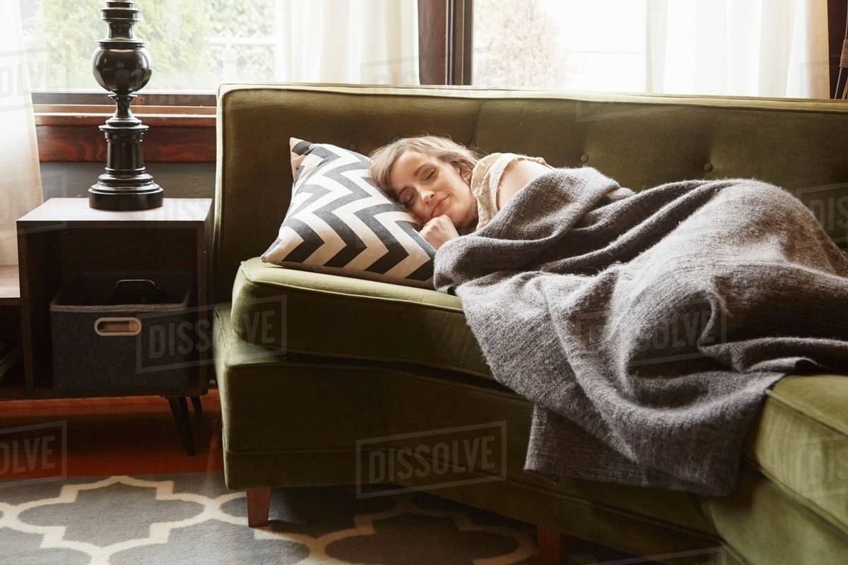 Young Woman Lying On Living Room Sofa Wrapped In Blanket Stock Young Woman Lying On Living Room Sofa Wrapped In Blanket