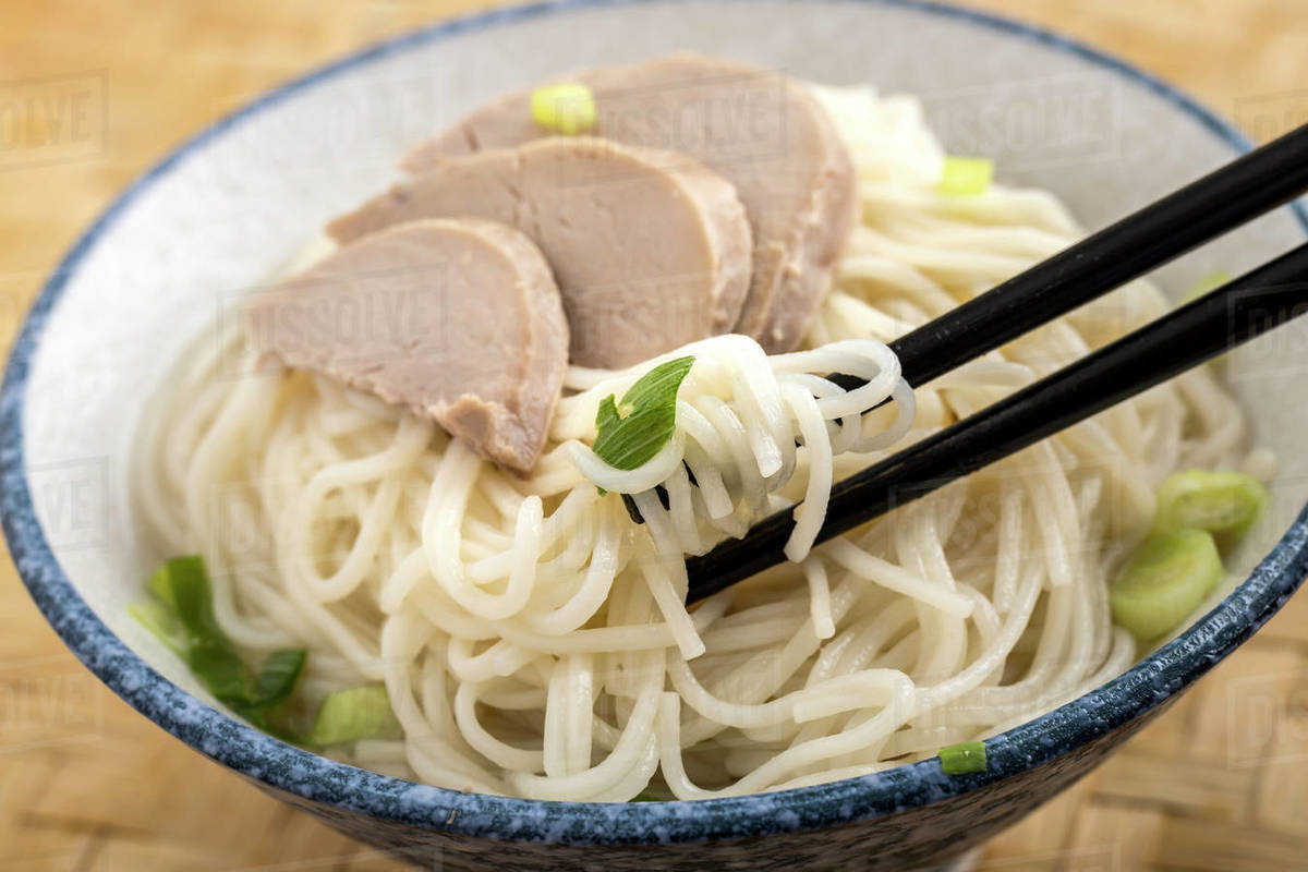 A close up photo of using chopsticks to eat a bowl of noodles topped with pork slices and green