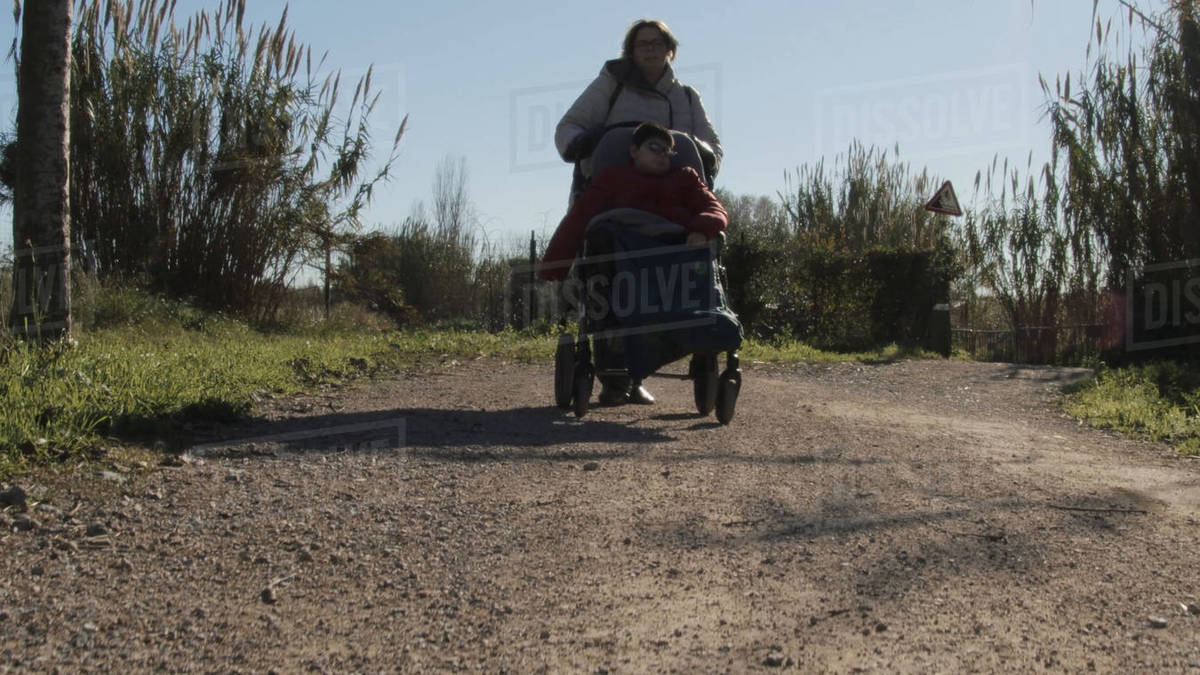 Woman wheelchair pass disabled child on a dirt road. Stock Photo