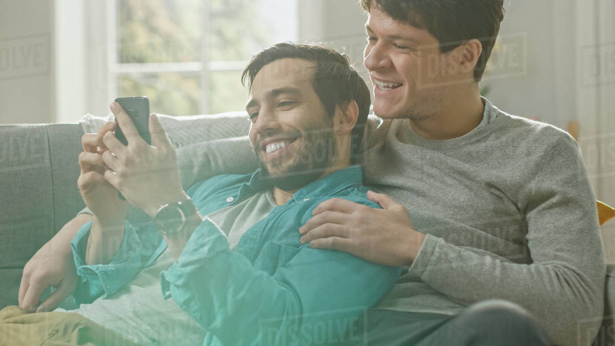 Cute male gay couple spend time at home. They are lying down on a sofa and use a smartphone. They browse online. Partner's hand is around his lover. They smile and laugh. Room has modern interior.  Royalty-free stock photo