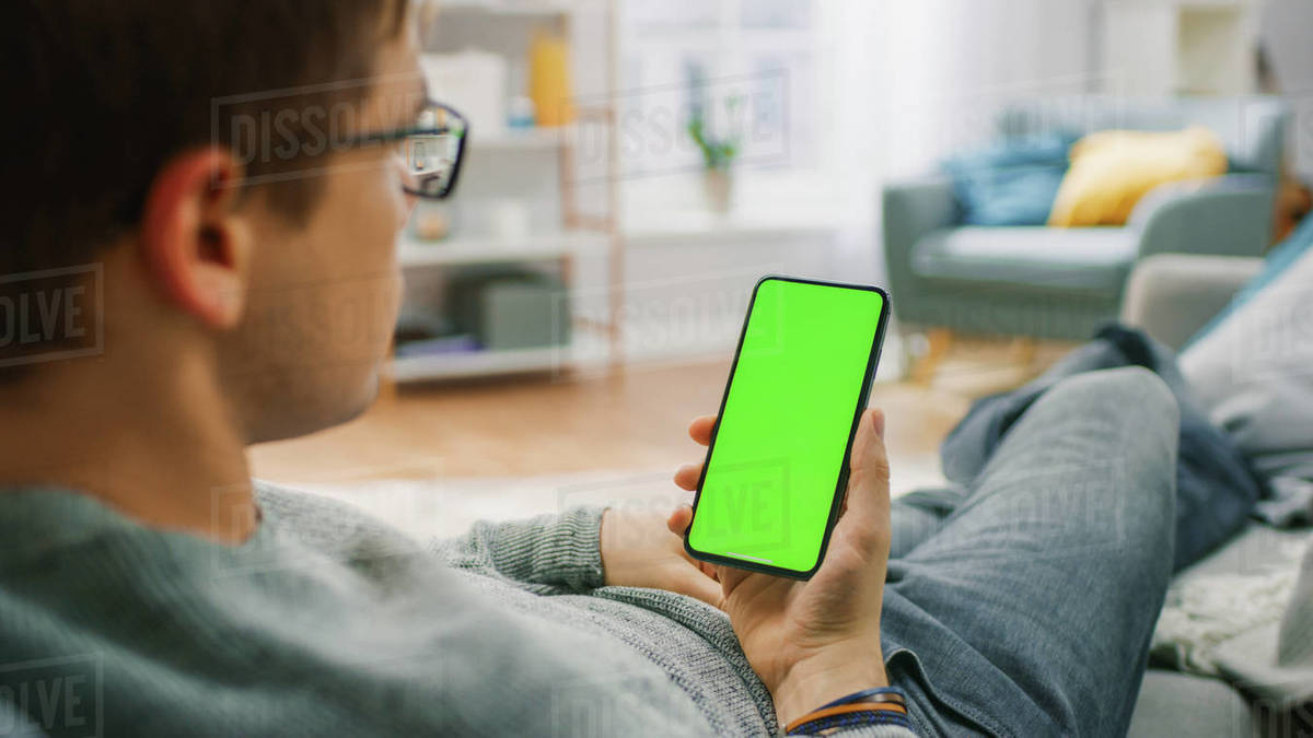 Man at Home Resting on a Couch using Smartphone with Green Mockup Screen. Guy Using Mobile