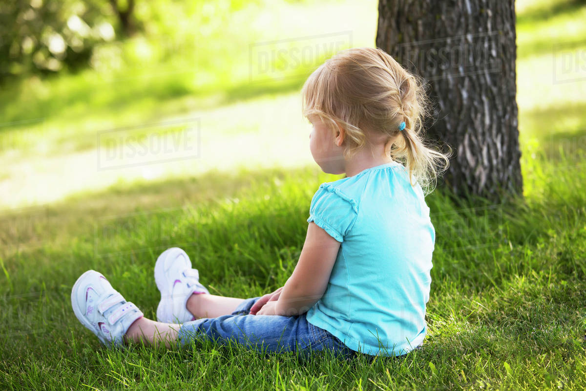 Toddler girl sitting in a park beside a tree; Edmonton, Alberta, Canada