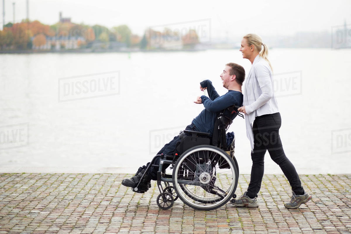 Caretaker pushing disabled man on wheelchair along lake - Royalty-free ...