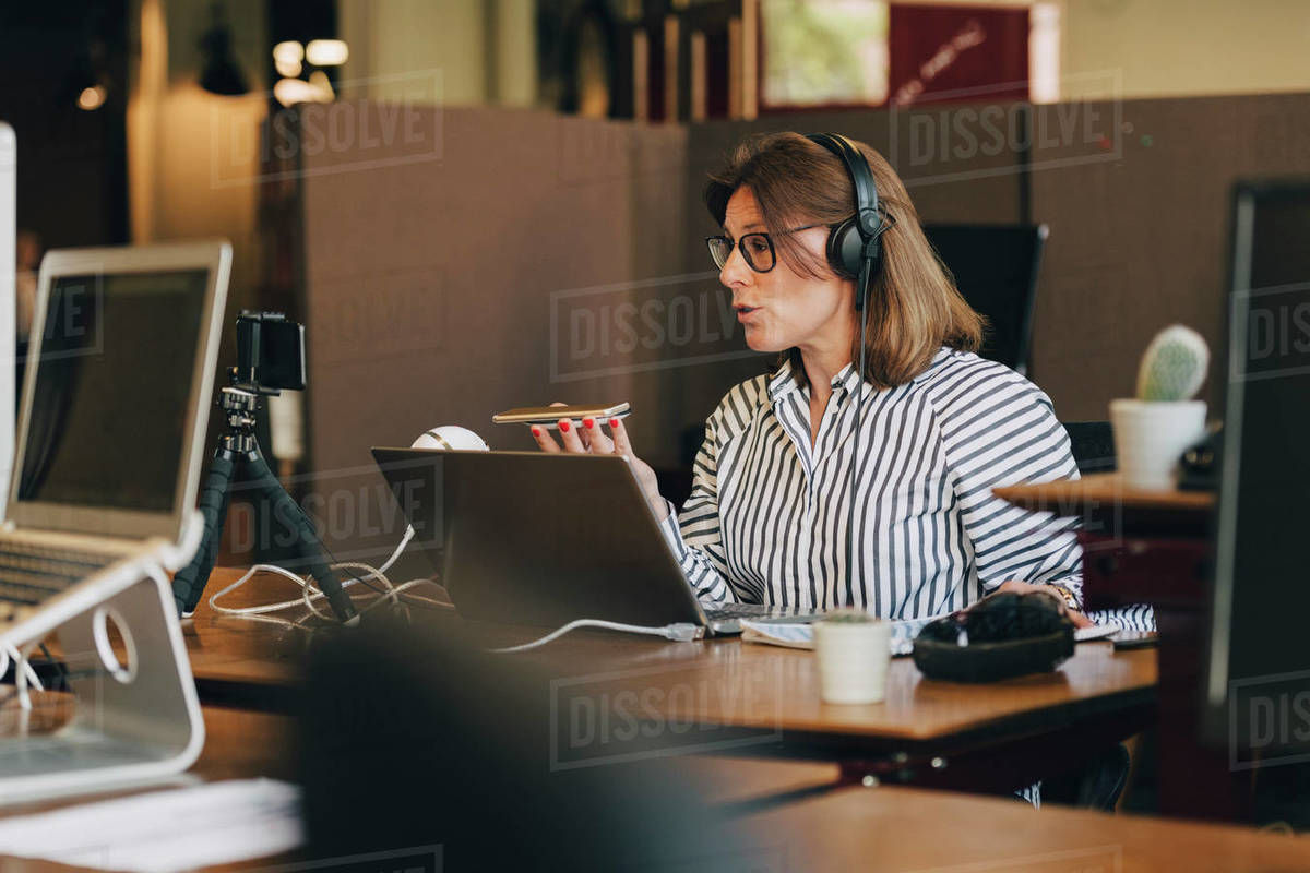 Mature businesswoman wearing headphones while talking on mobile phone in office Royalty-free stock photo