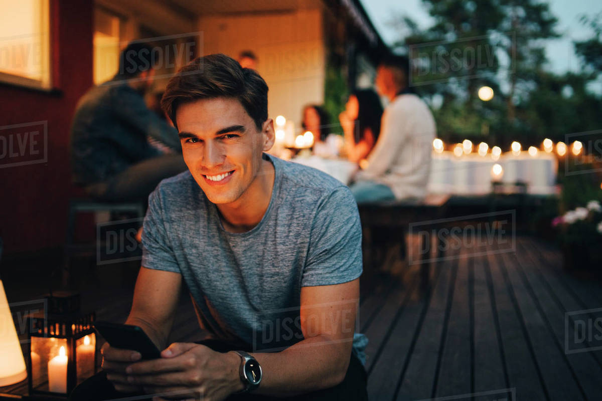 Portrait of smiling young man holding smart phone while friends in background during dinner party Royalty-free stock photo