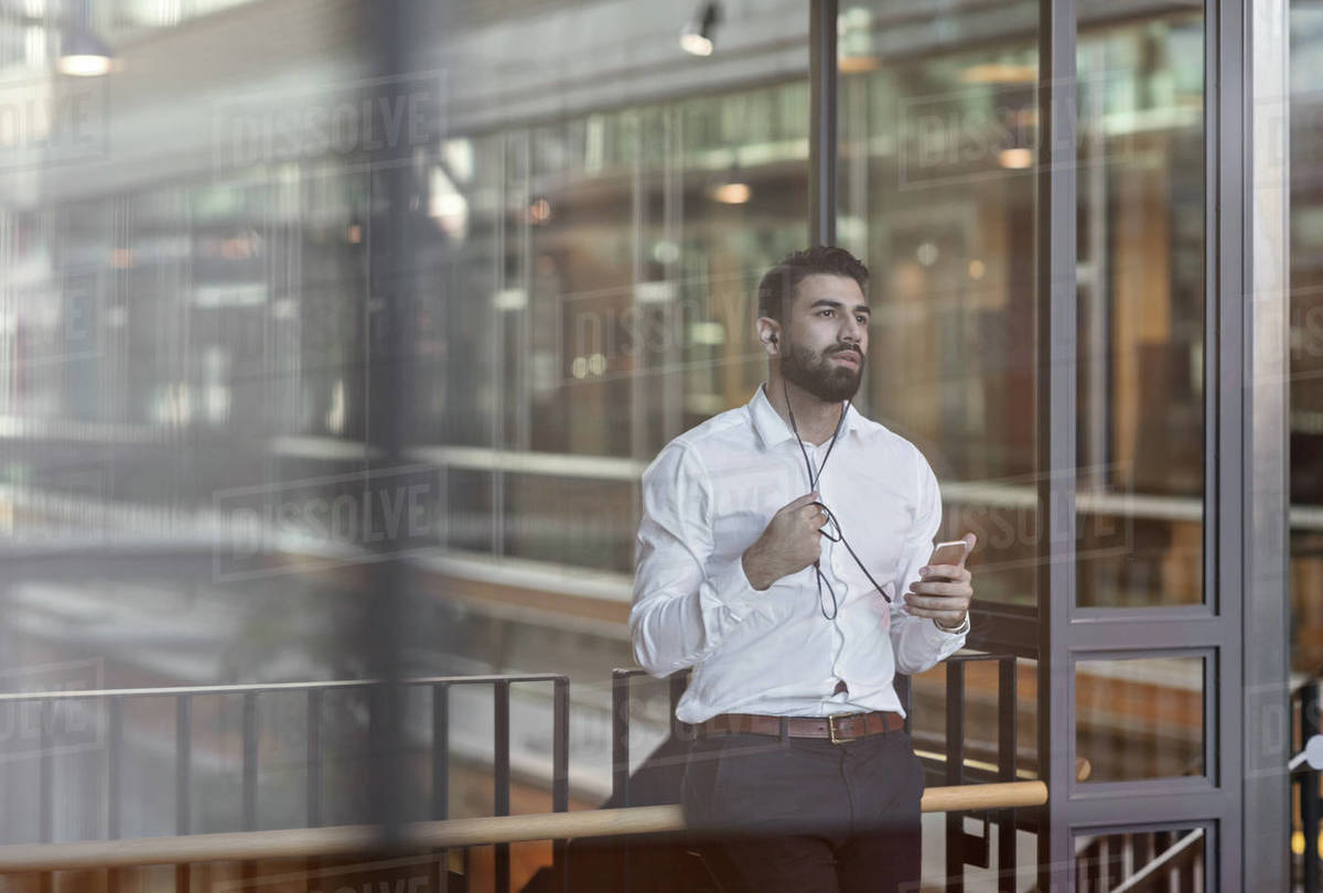 Businessman wearing headphones and using smart phone while standing in lobby at office Royalty-free stock photo