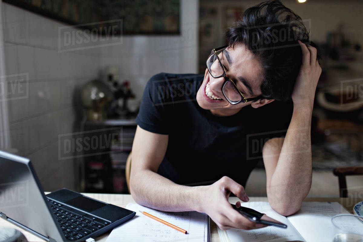 Cheerful social media addicted young man using mobile phone while doing homework Royalty-free stock photo