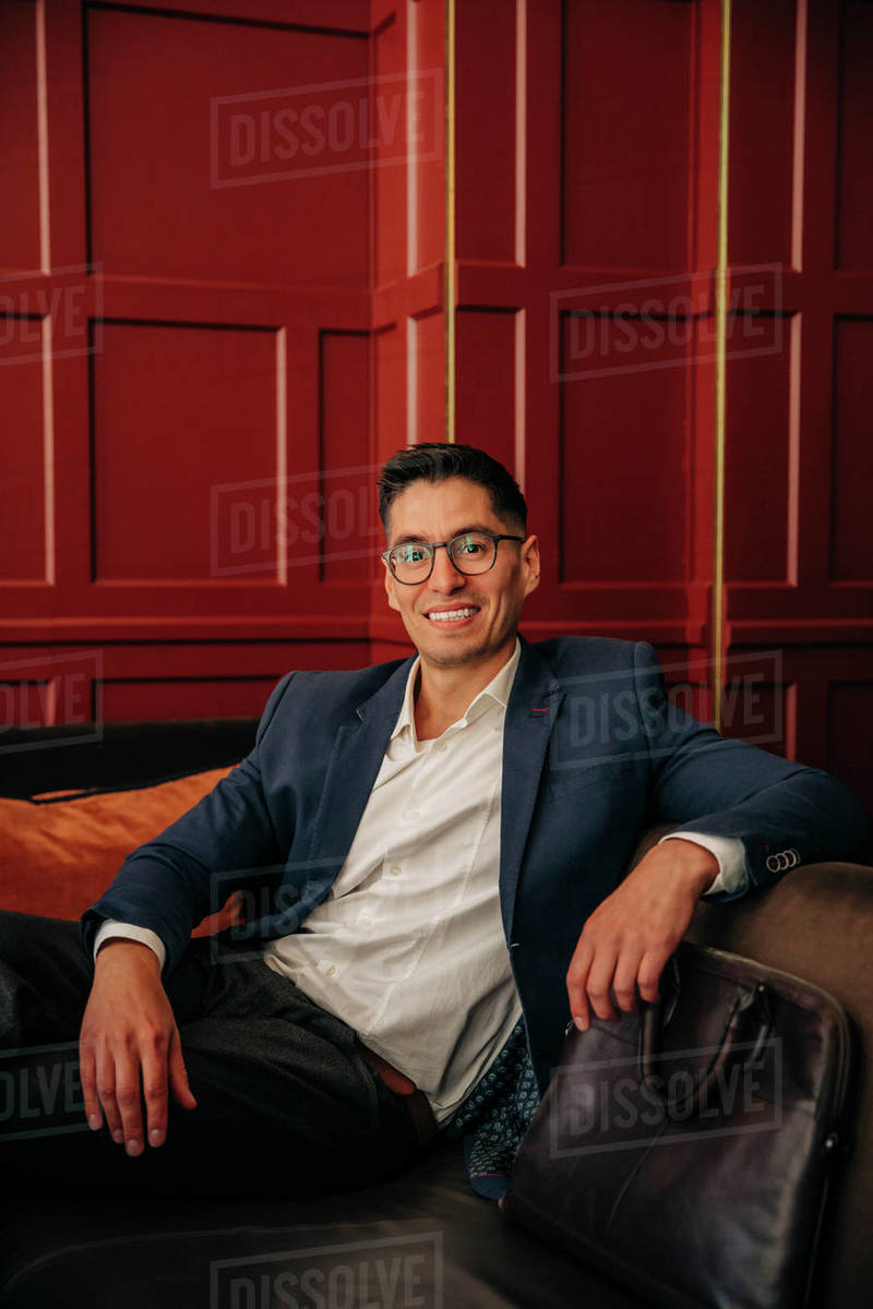 Happy male entrepreneur wearing eyeglasses sitting on sofa in hotel lounge Stock Photo Dissolve