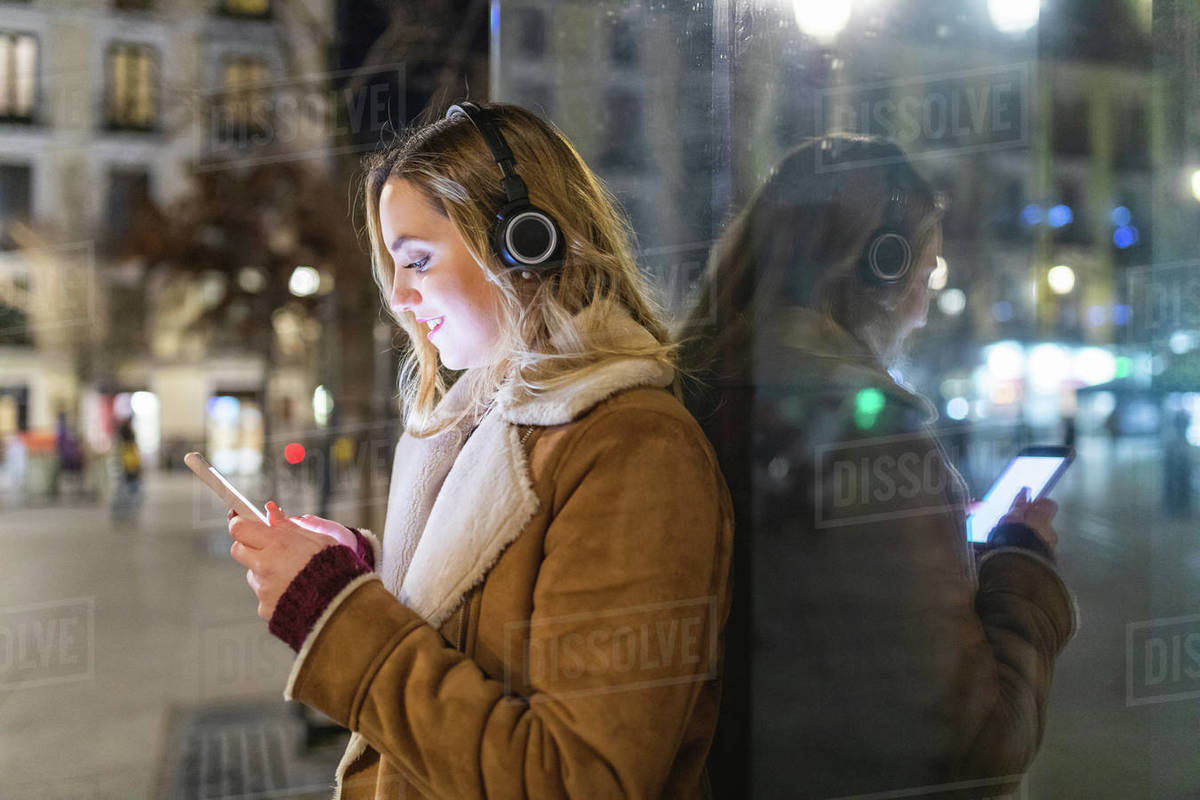 Young woman leaning against shop window listening to headphones and looking at smartphone at night, Madrid, Spain Royalty-free stock photo