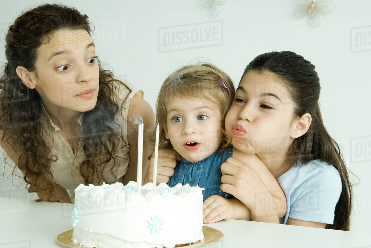 Girl helping younger sister blow out candles on birthday cake, mother
