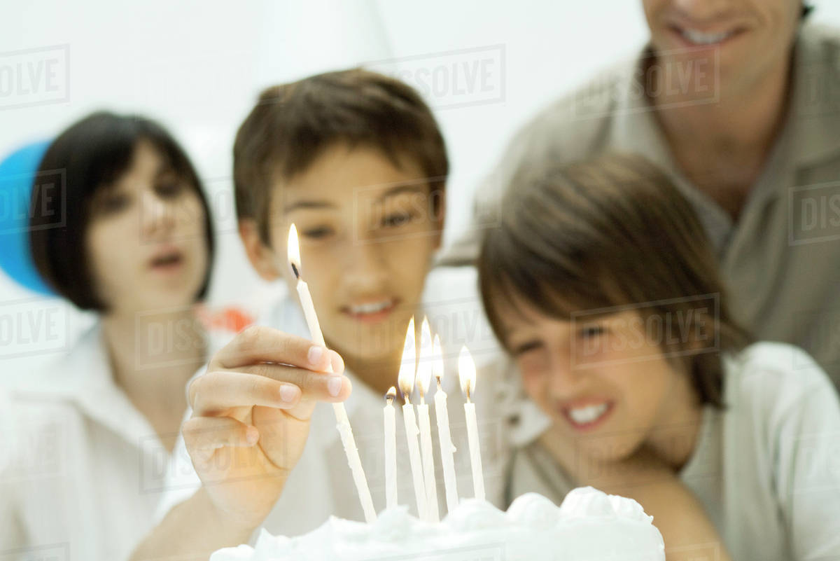 Boy placing burning candles on birthday cake, family watching Stock
