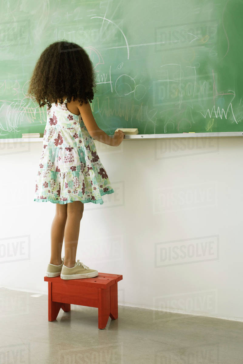 Little girl standing on stool in front of blackboard, rear view Stock Photo Dissolve