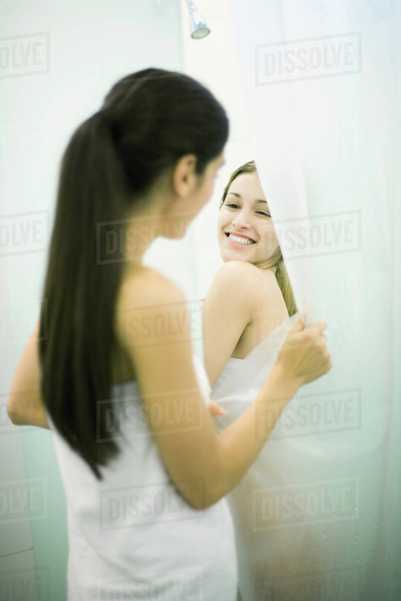 Young woman taking shower while friend holds open shower curtain