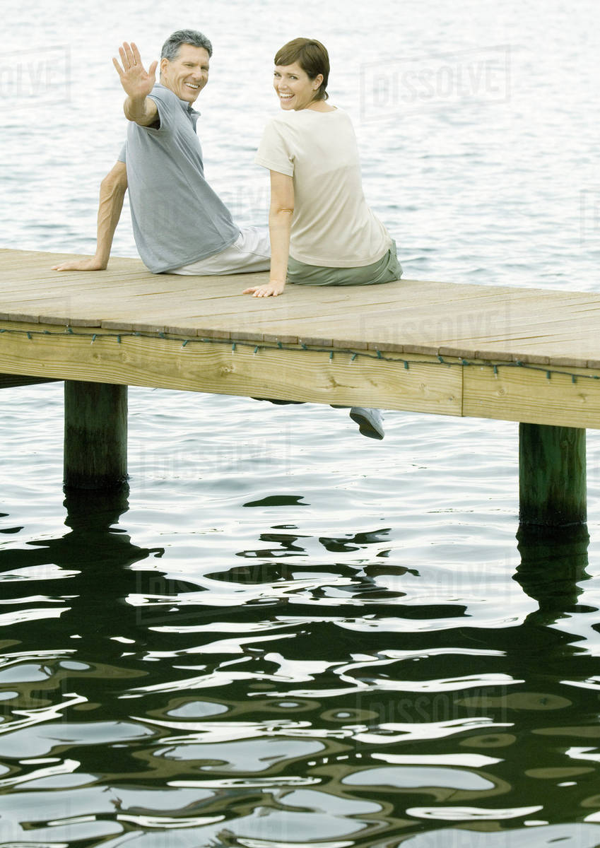 Couple sitting on dock, man waving at camera Stock Photo Dissolve