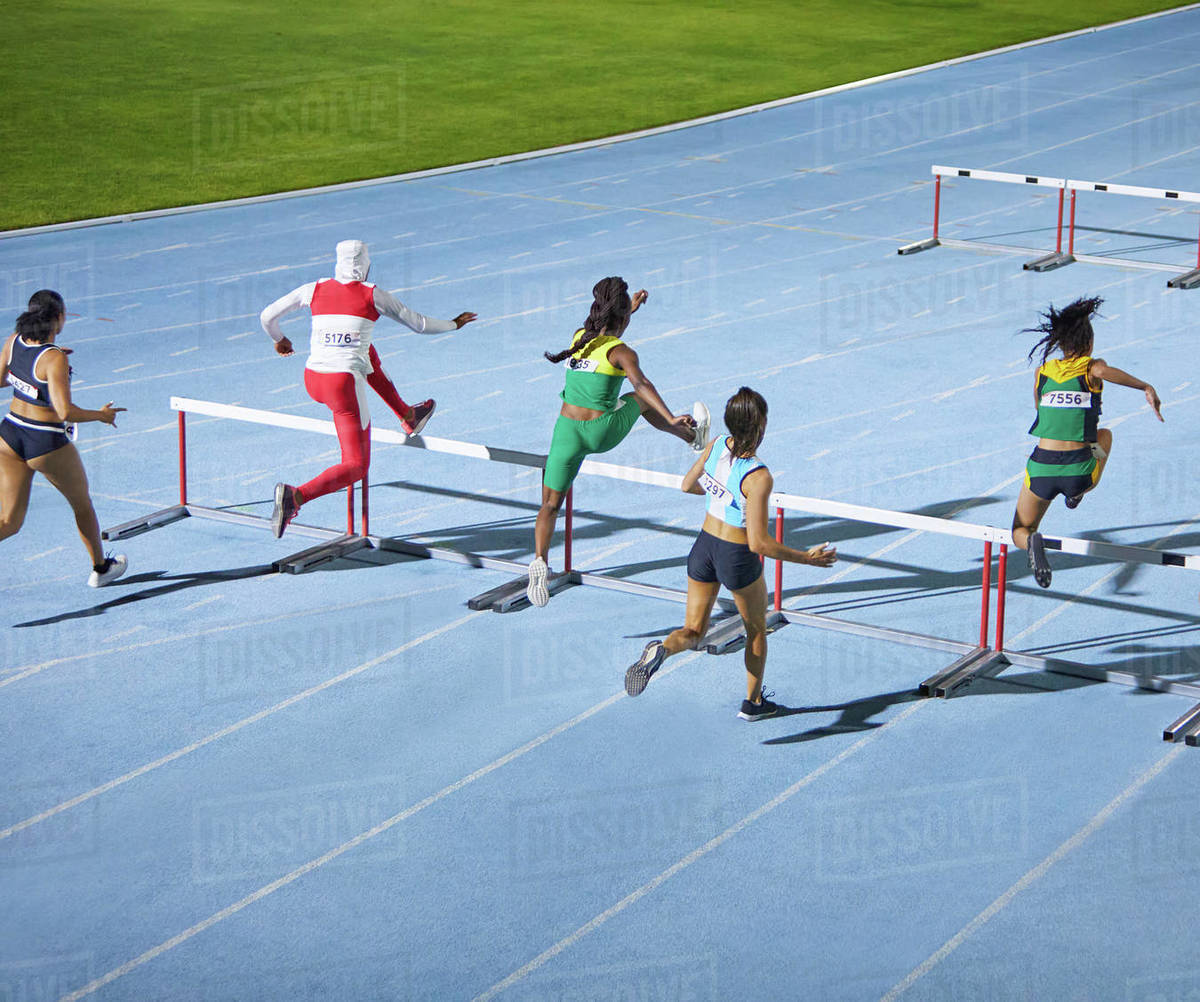 Female track and field athletes jumping hurdles in competition Stock